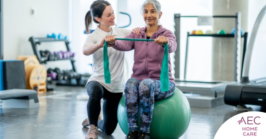A care provider helps an older woman exercise with a resistance band and an exercise ball, representing how exercise can help with senior fall prevention.
