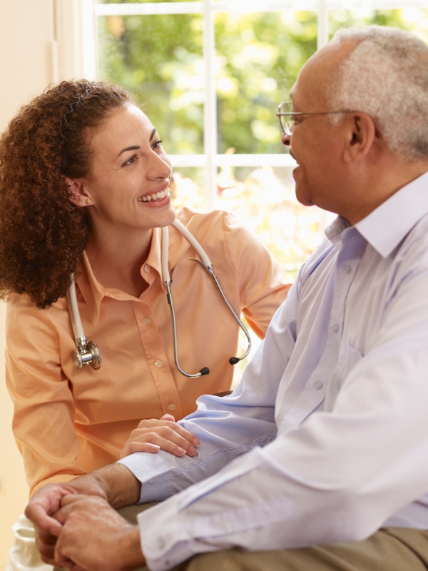 A nurse with a stethoscope smiles while speaking with an elderly man during an in-home visit, offering comfort, companionship, and attentive care.