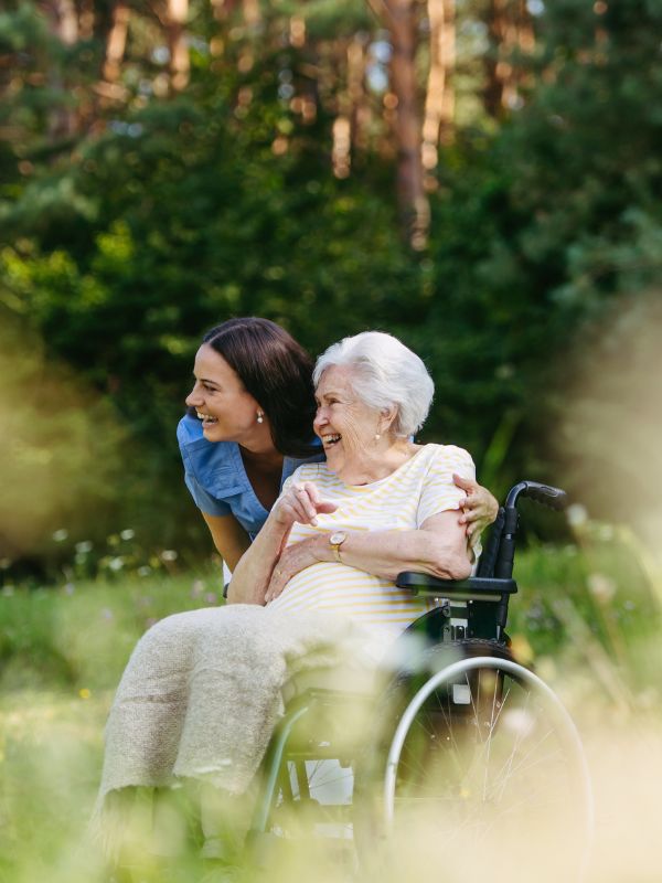 Caregiver Assisting Senior Woman Outdoors A smiling caregiver leans beside an elderly woman in a wheelchair while they enjoy time together outdoors in a sunny green park.