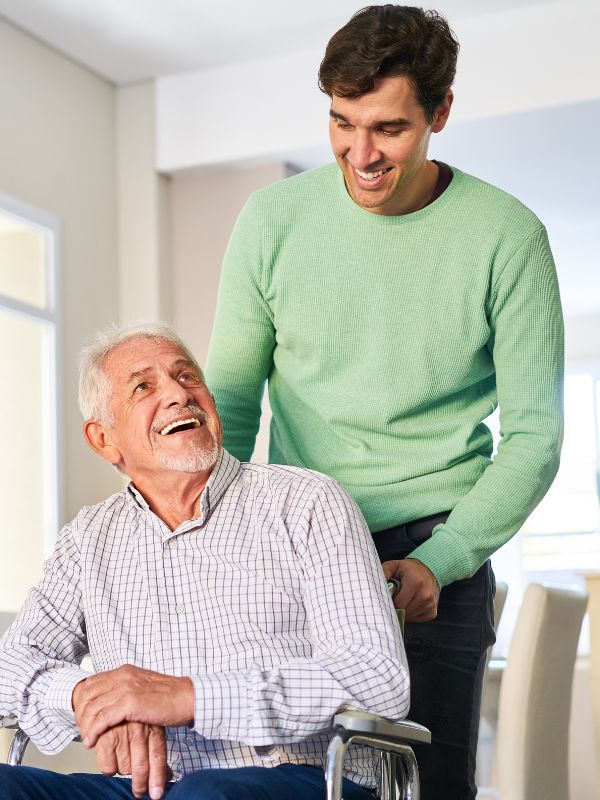 A smiling caregiver stands behind a senior man in a wheelchair, both sharing a warm, cheerful moment inside a bright home.